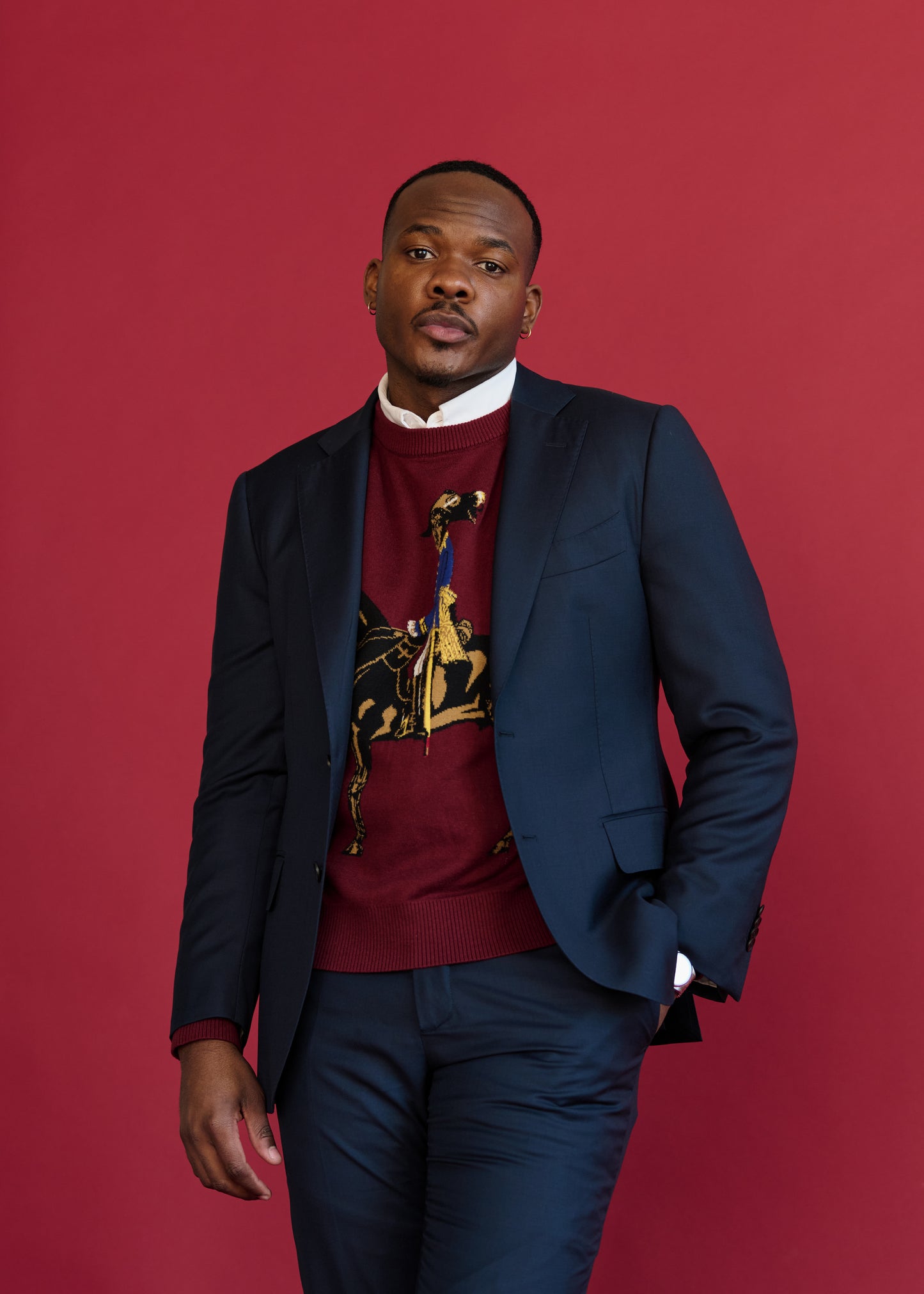 Designer and model wearing the Toussaint Sweater in burgundy, styled with a navy suit jacket in front of a red backdrop